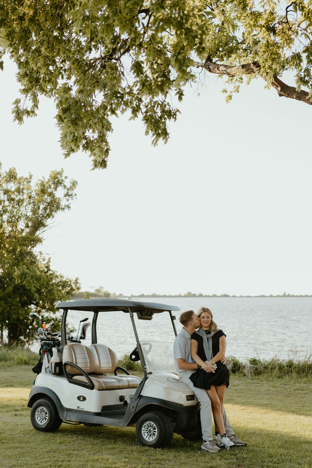 Couple kissing beside a white golf cart near a serene waterfront at sunset.