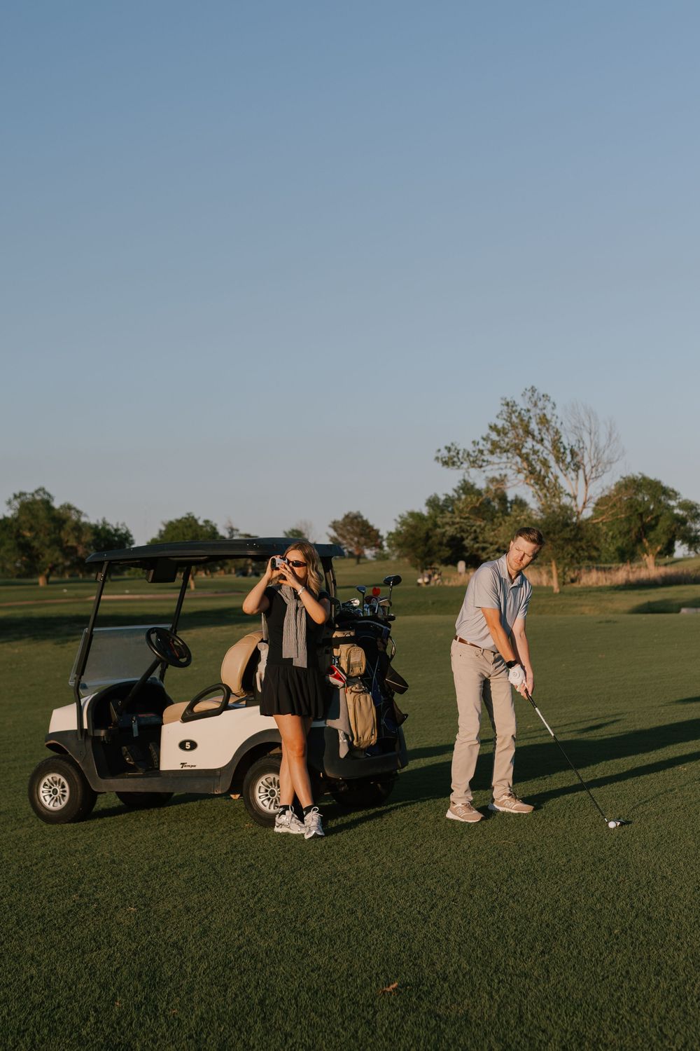 Two golfers on a sunny golf course, one preparing to putt while the other takes a photo near a white golf cart.