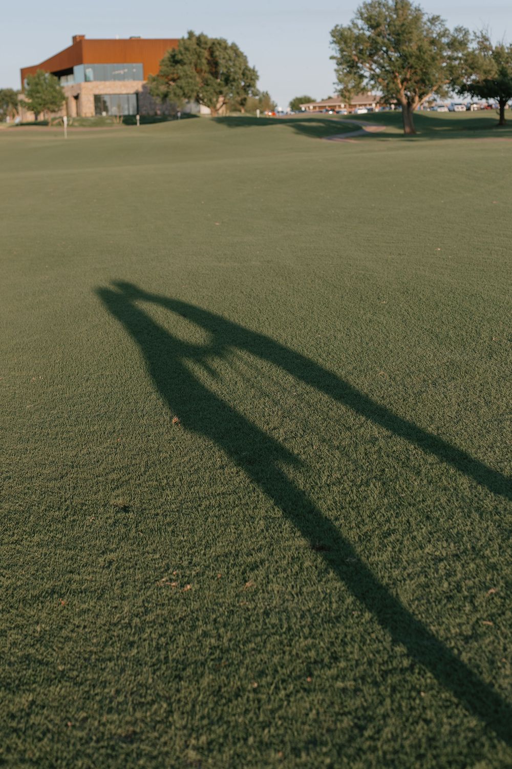 Shadow of a golfer cast on a lush green golf course with trees and a clubhouse in the background.