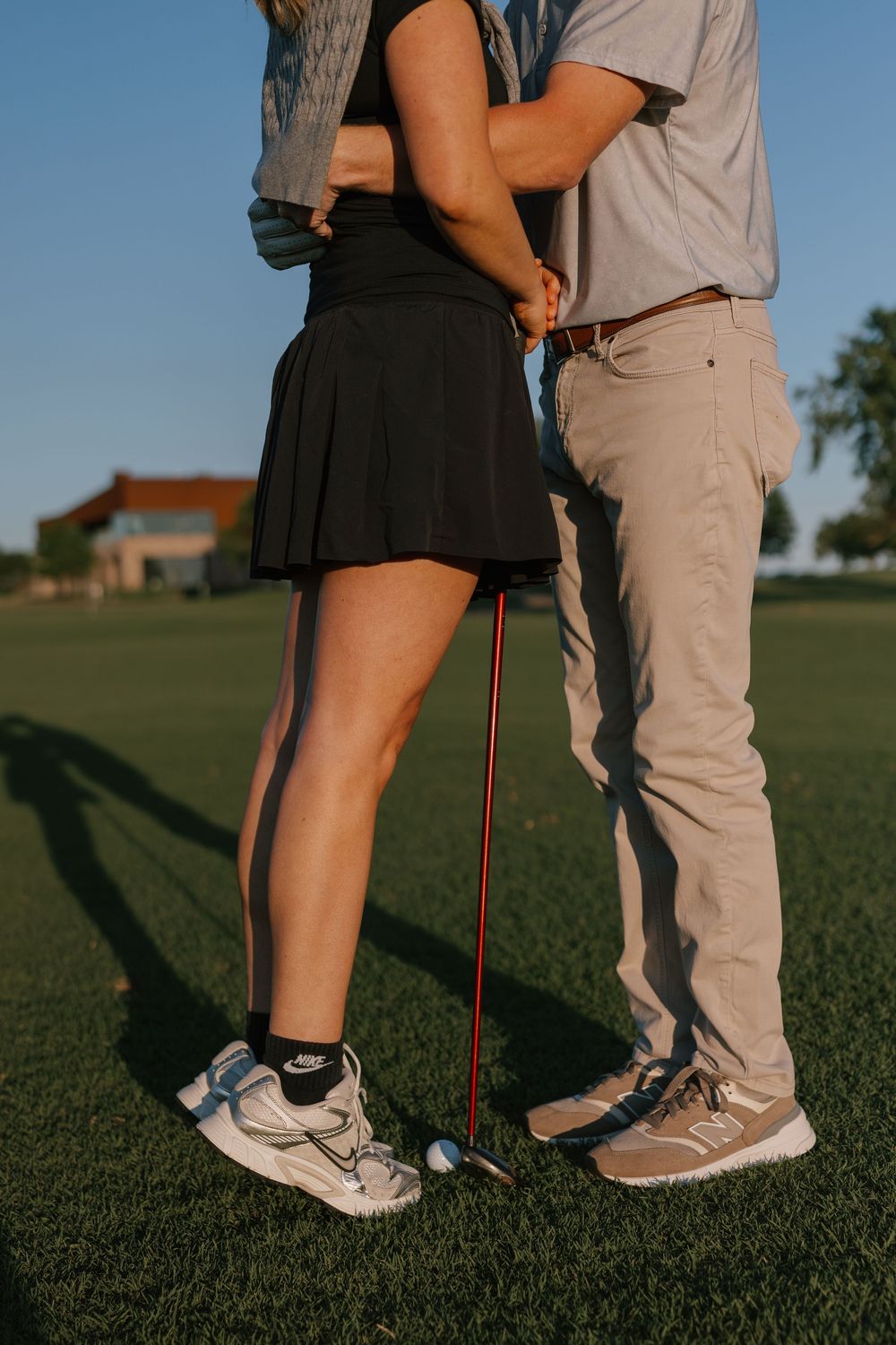 Couple standing close together on a golf course, holding a golf club, wearing casual golf attire at golden hour.