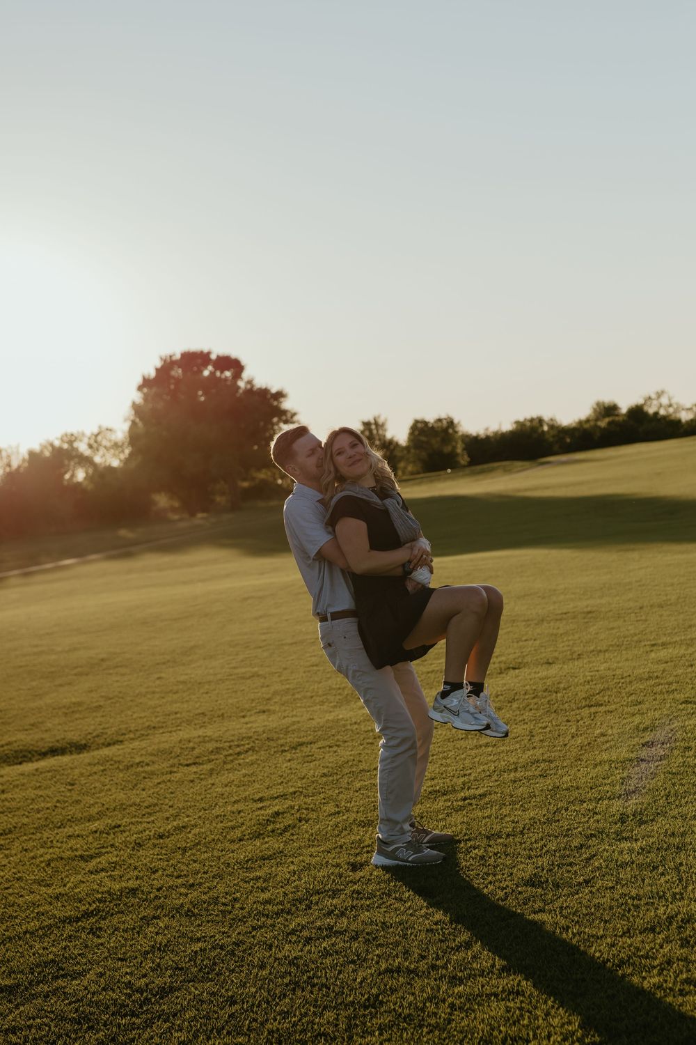 Man lifting woman in his arms on a sunlit golf course at golden hour.