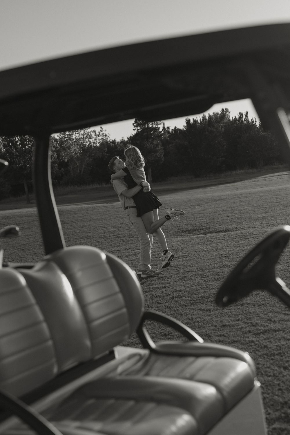 Black and white photo of couple embracing on golf course viewed through cart.