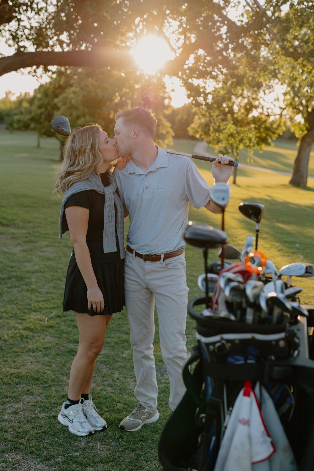 Couple sharing a kiss at sunset on a golf course near a bag of golf clubs with warm golden backlight.