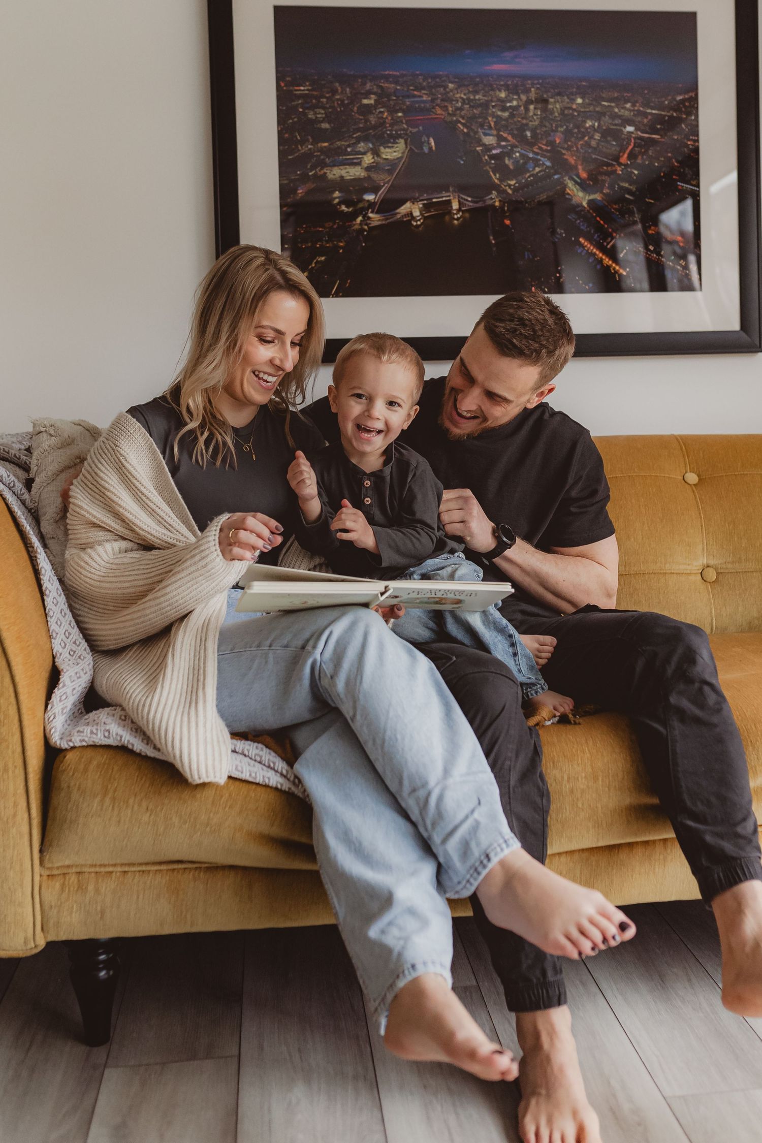 Happy family of three sitting on a yellow sofa, smiling while looking at a photo album together in a cozy living room.