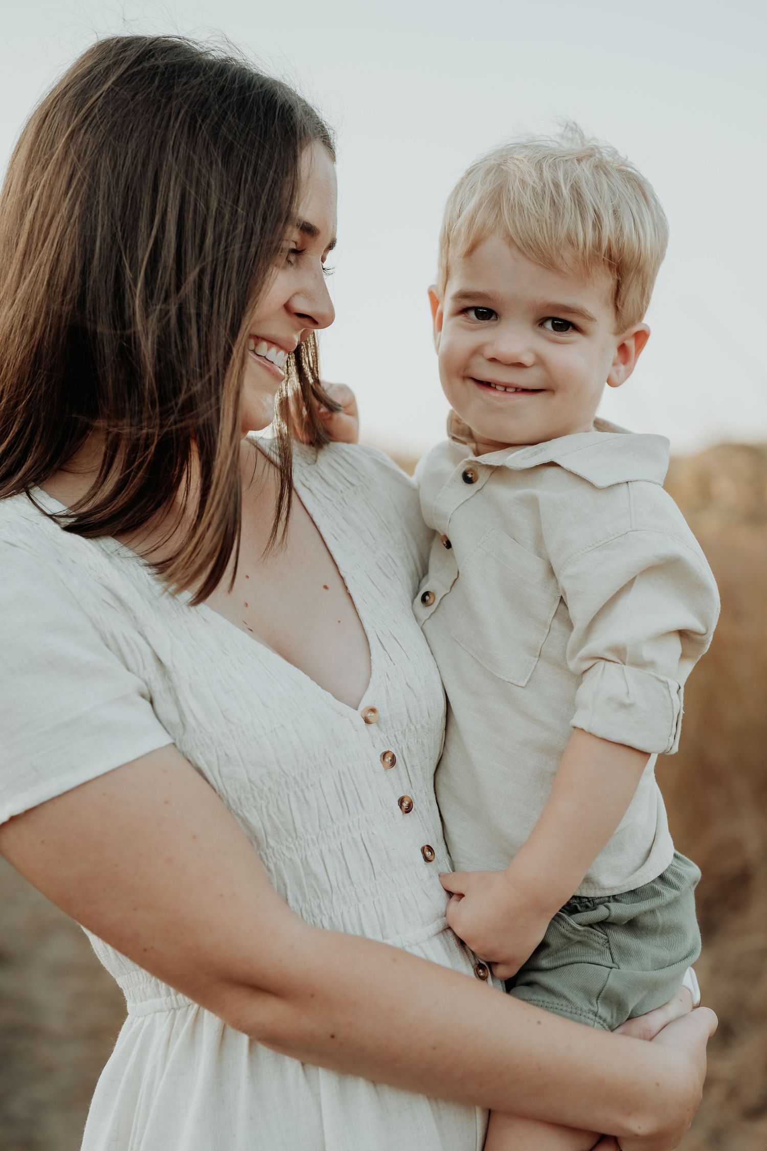 A smiling mother in a white dress holds her happy blonde toddler boy outdoors in a golden field.