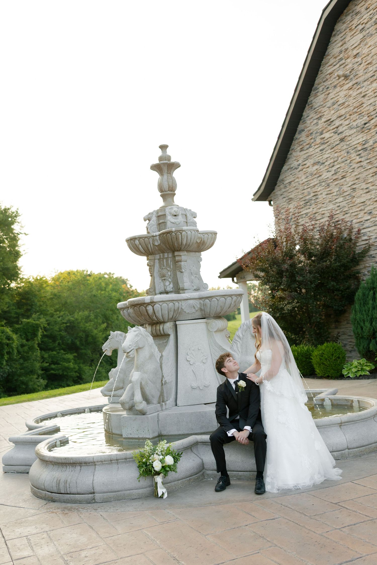 Bride and groom share a romantic moment beside an elegant stone fountain at sunset.