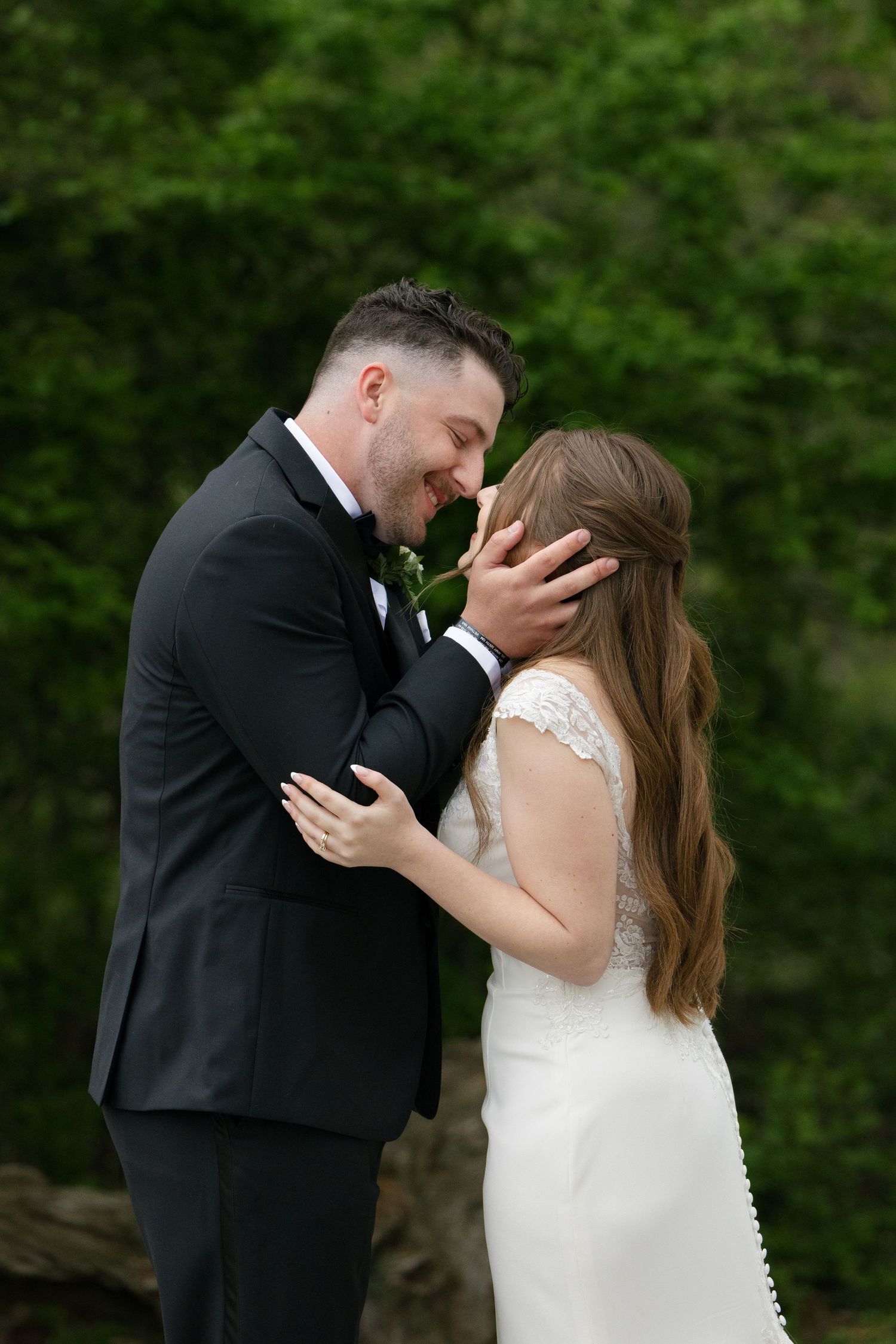 Groom tenderly holds bride's face as they share an intimate forehead touch outdoors.