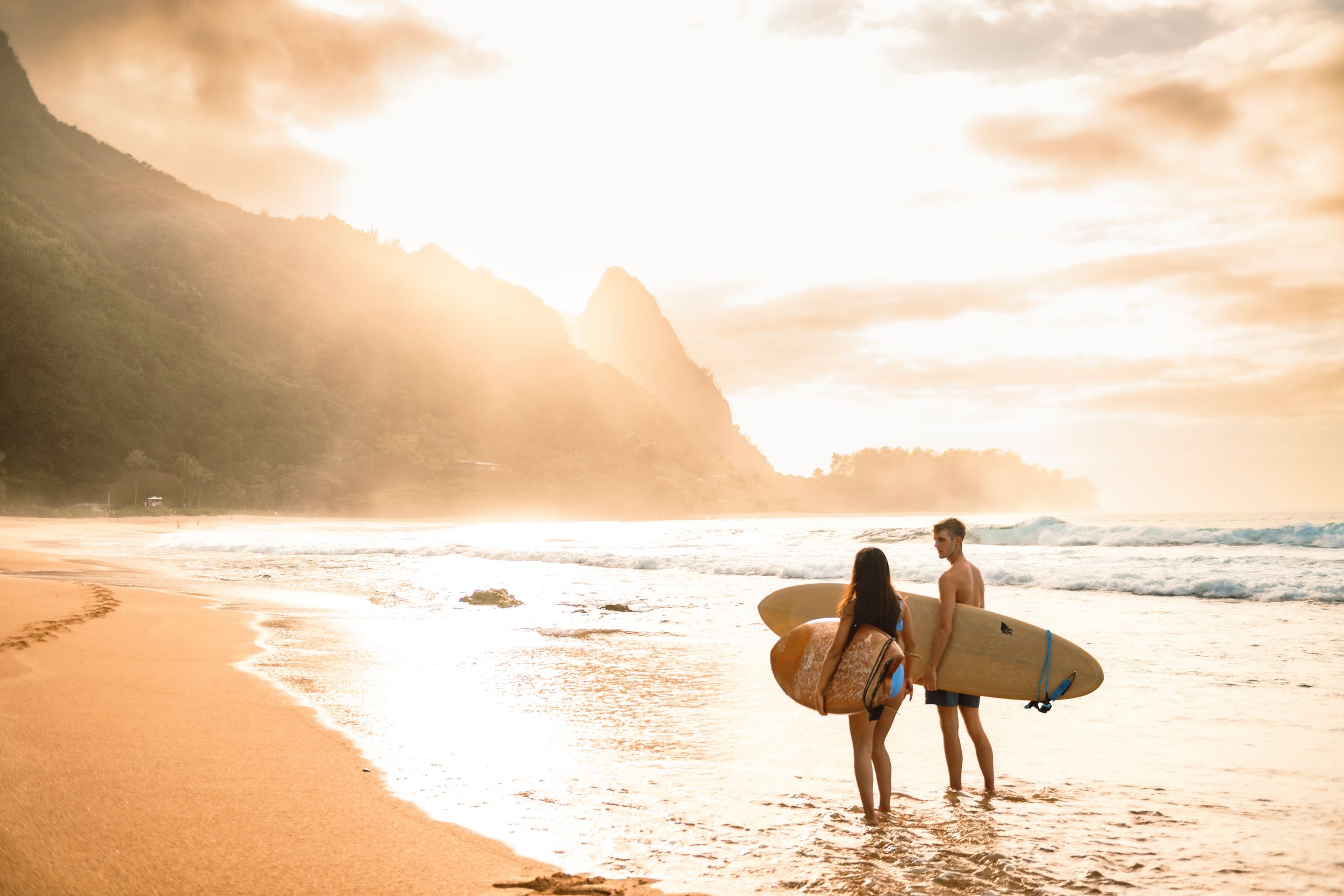 Two surfers holding boards stand on Maui sandy beach in Hawaii at sunset with misty mountains in the background.