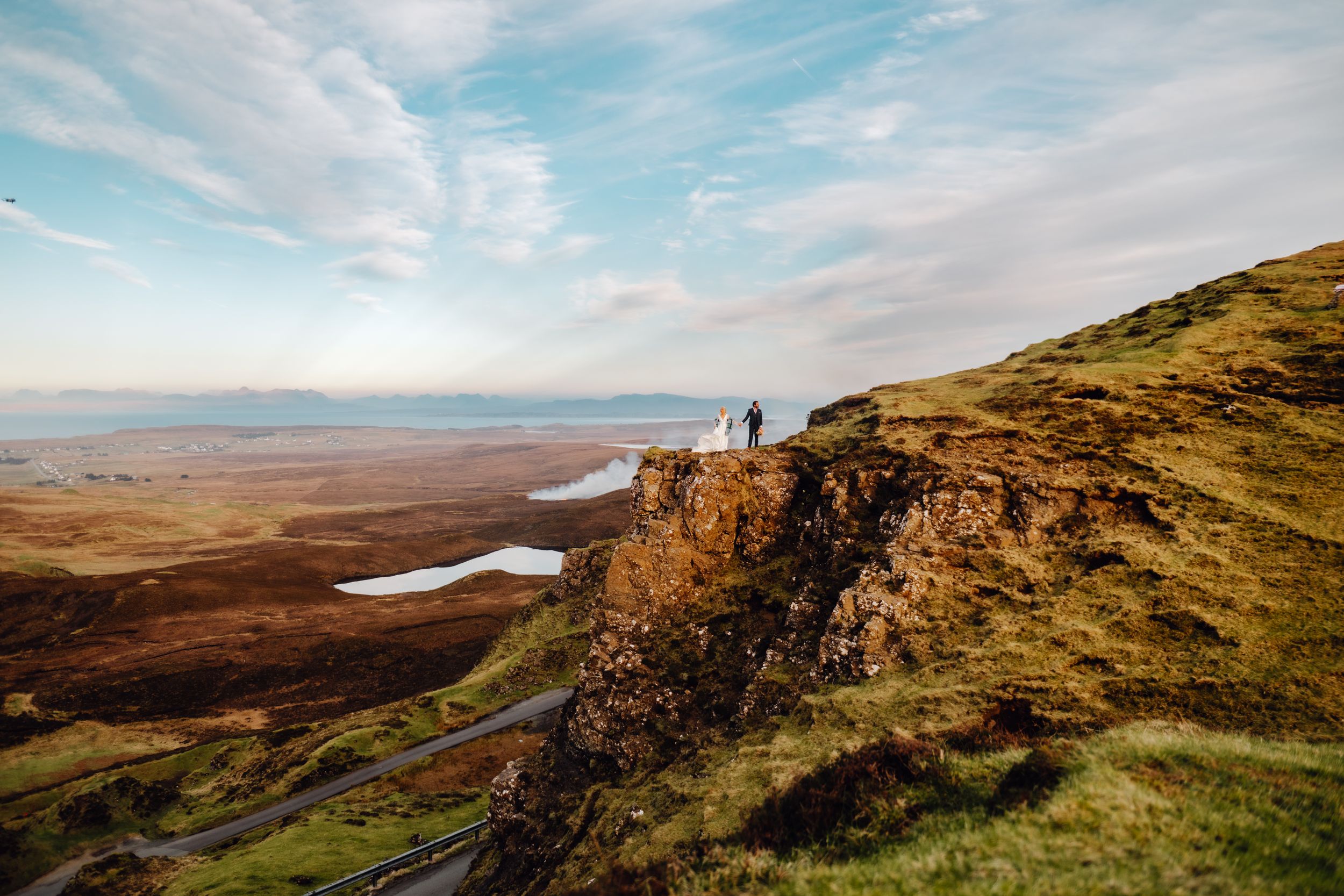 Couple stands on dramatic Scottish Highland cliffside with sweeping valley and loch views below.