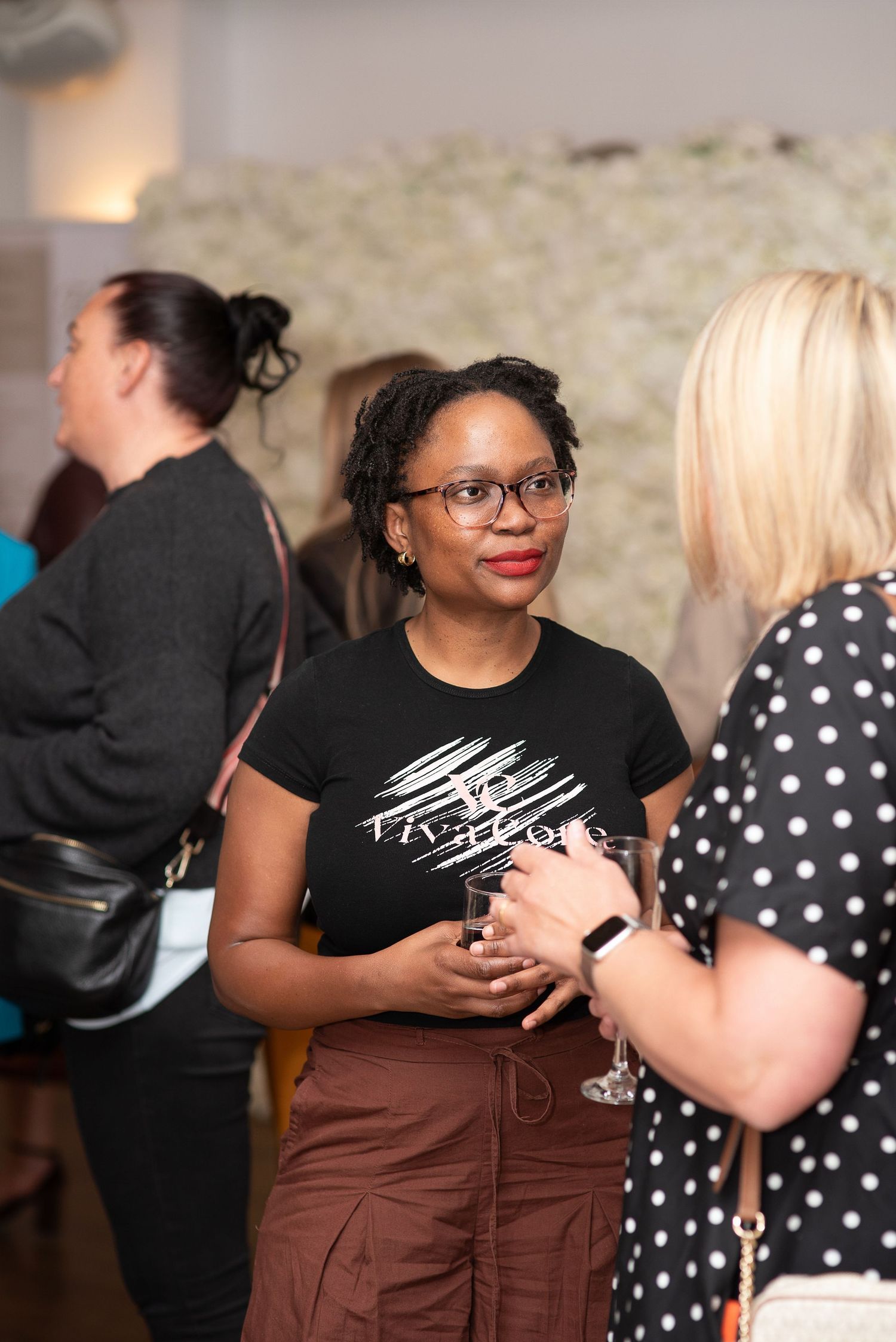 Women networking at a social event, smiling and holding drinks in a stylish venue with floral backdrop.