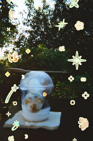 A decorative ceramic jar with angelic figures sits outdoors, surrounded by digital star and flower sticker overlays.