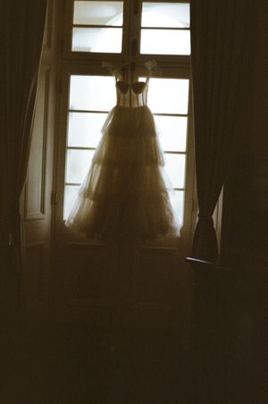 A elegant white wedding dress hanging in a window, backlit by natural light in a dark romantic room.
