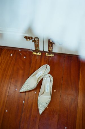 Elegant gold glitter Jimmy Choo bridal heels on hardwood floor beside white wedding dress and gold door hardware.