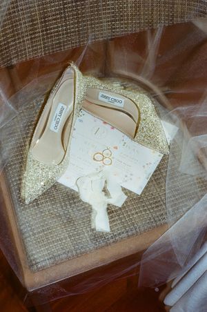 Gold glitter Jimmy Choo bridal heels displayed with wedding rings and invitation on a textured surface beneath sheer veil.
