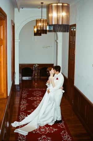 Bride and groom share a romantic kiss in an elegant hallway with red carpet and vintage chandeliers.