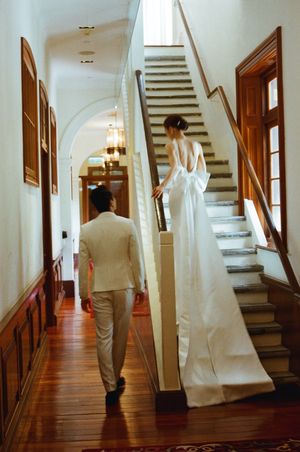 Bride in backless white gown ascending staircase as groom follows in elegant historic venue hallway.