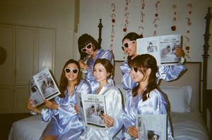 Five friends in matching blue satin robes and white sunglasses pose on a bed reading newspapers in a decorated room.