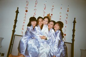 Five women in matching silver satin robes pose together smiling on a bed with decorative streamers in the background.