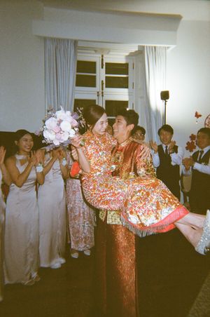 Couple in traditional Chinese wedding attire, groom carrying bride while guests celebrate with bouquet toss indoors.