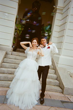 Joyful couple in wedding attire posing on steps, bride in white tulle gown, groom in white tuxedo jacket.