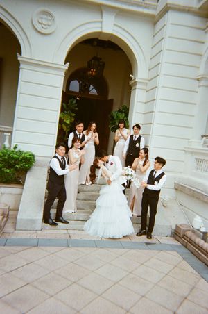 Wedding party poses on grand white building steps, bride in white gown with bouquet surrounded by bridesmaids and groomsmen.