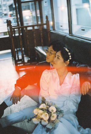 Bride and groom in wedding attire sit together on a red seat inside a vintage tram, bride holding a bouquet.