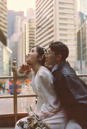 Bride and groom in wedding attire gaze upward on a tram balcony with city skyscrapers in the background.