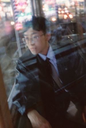 A young man in glasses and suit seen through a blurred window reflection with city lights at night.