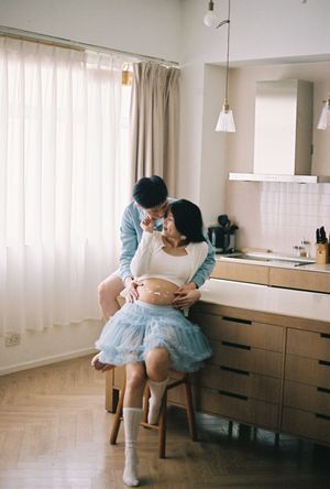 A couple shares a tender moment in a cozy kitchen, with the pregnant woman seated on a stool wearing a blue tulle skirt.