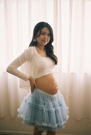 Pregnant woman in white top and blue tutu skirt posing with baby bump decorated with stickers.