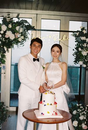 Bride and groom cutting a two-tier white wedding cake decorated with colorful flowers beneath a floral arch.