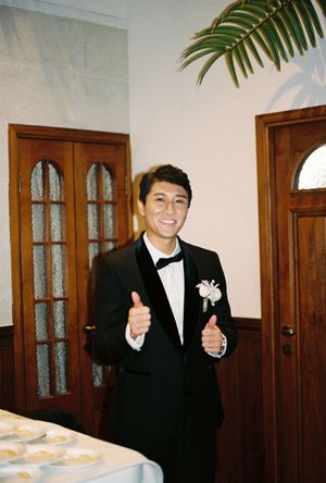 Young man in black tuxedo with boutonniere giving thumbs up and smiling at formal event.