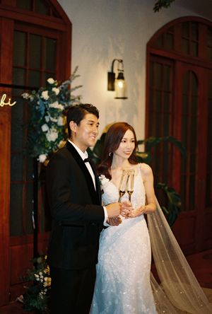 A bride and groom toast with champagne glasses at their elegant wedding reception, she in a lace gown with veil, he in a black tuxedo.
