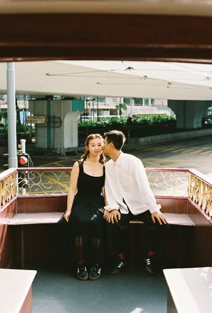 A couple sits closely together on a decorative boat bench, the woman in a black dress and man in white shirt, in an urban setting.