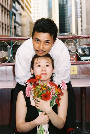 Couple posing with colorful flower bouquet on urban balcony, man standing behind seated woman in black dress.