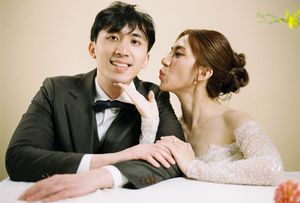 A bride in a lace gown kisses groom's cheek as he smiles in a black tuxedo during wedding photo session.