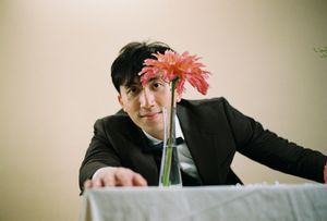A smiling man in a black suit leans toward camera beside a glass vase with a pink gerbera daisy flower.