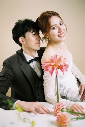 A couple in wedding attire poses with pink flowers, the groom in a dark suit and bride in a beaded white gown, smiling warmly.