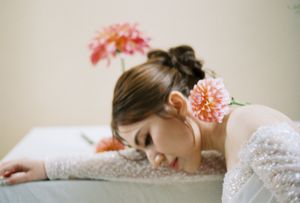 Young woman in white lace dress resting head on table, adorned with pink dahlia flowers in her hair.