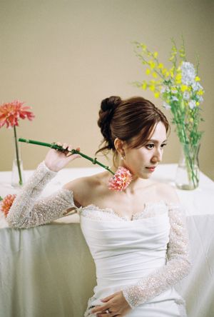 Bride in white off-shoulder gown holds pink dahlia stem, seated near floral arrangements in soft neutral setting.