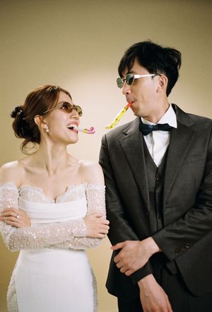A bride and groom in sunglasses playfully blow party favors at each other, smiling at their wedding celebration.