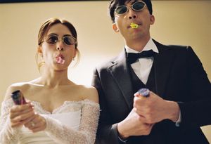 Bride and groom in sunglasses blowing party poppers at their wedding reception, wearing formal attire.