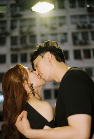 A couple shares a romantic kiss at night under a bright light with a building facade in the background.