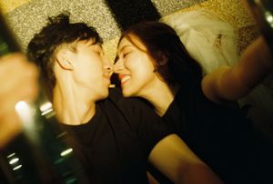 A young couple lying close together, sharing an intimate moment, smiling at each other in a dimly lit room.