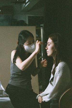 A makeup artist wearing a face mask carefully applies makeup to a seated woman in a dimly lit studio.