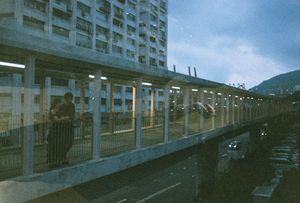 Elevated pedestrian walkway alongside a high-rise building at dusk, with city traffic below and mountains in the background.