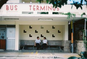 Two people sitting at a retro Bus Terminus shelter, minimalist architecture with decorative wall patterns.