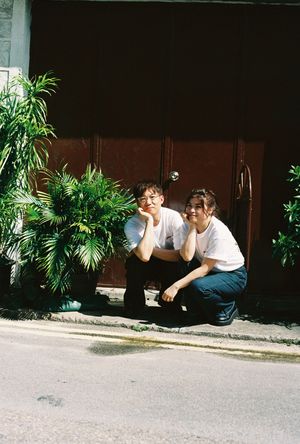 Two young people in white t-shirts squat smiling beside lush green plants outside a building on a sunny day.