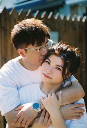 A young couple in white t-shirts embrace outdoors, man with glasses hugging woman from behind near wooden fence.