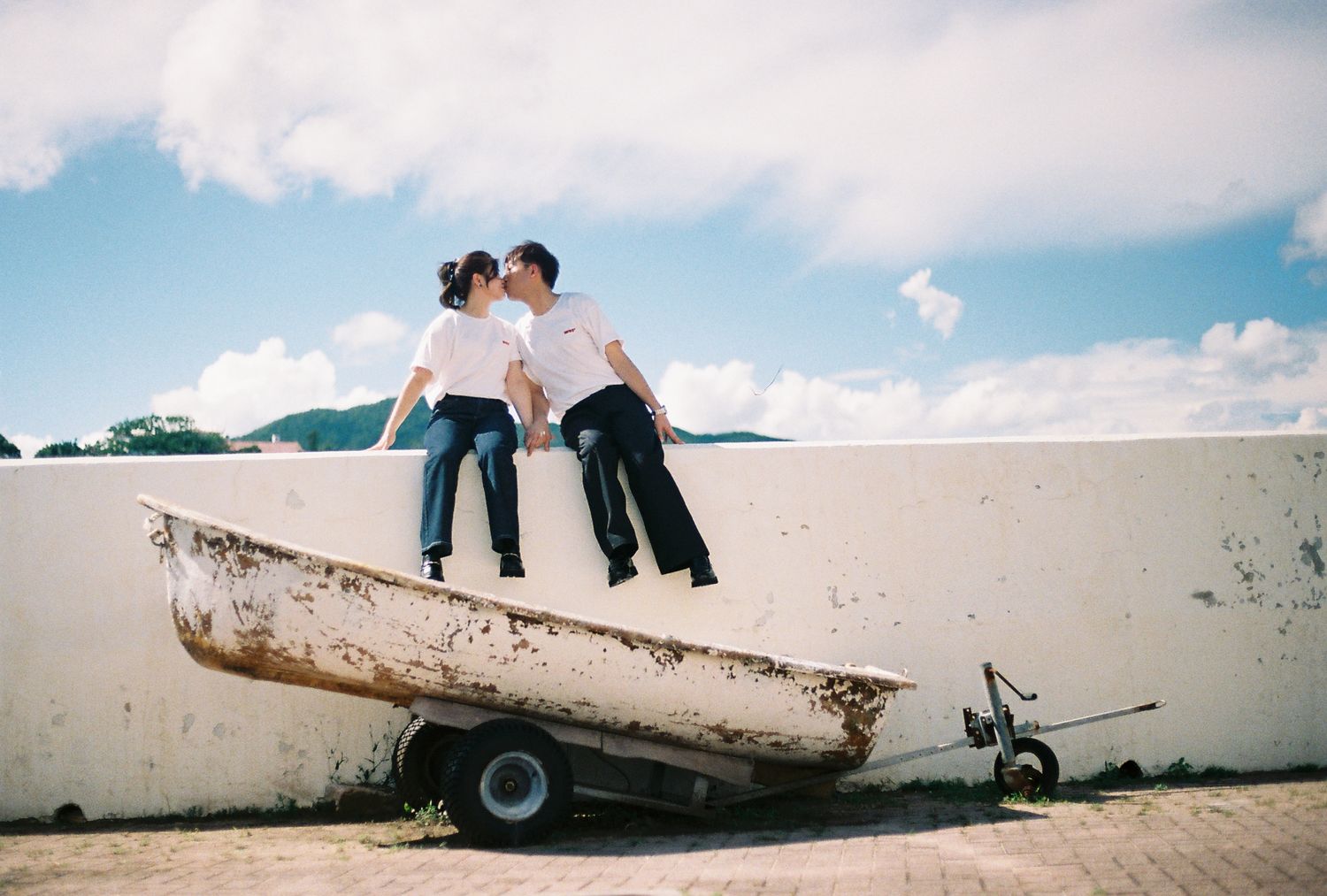 Couple kissing while sitting on a weathered boat on a trailer against a blue sky backdrop.